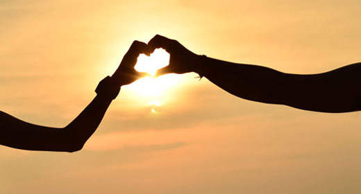 Two people making a hear symbol with their fingers in the setting sun.