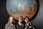 Three women posing in front of a large realistic moon installation